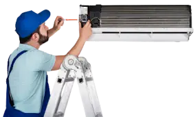 A man wearing a Sky Bulle shirt, blue pants & blue cap, climbs on a rack and repairs an air conditioner