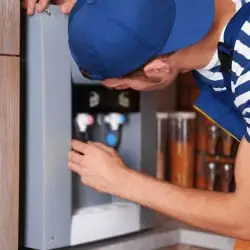 A man is wearing a blue & white checks t-shirt and is inspecting a water purifier for repairs